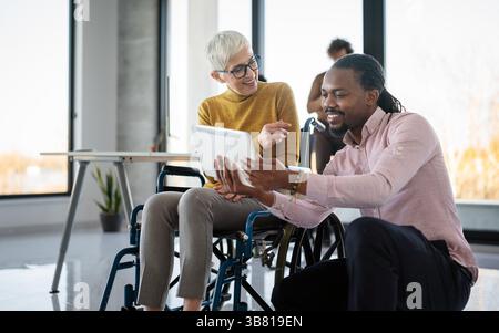 Disabled woman in wheelchair looking at computer, discussing startup project with interracial team Stock Photo