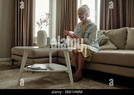 Senior Woman Seated on Couch Checking Device in Living Room Stock Photo