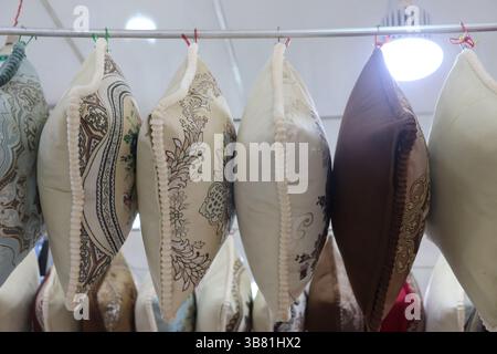 Assorted decorative cushions with unique patterned fabrics and braided trim hang from a metal rod, illuminated by an overhead light in a retail enviro Stock Photo