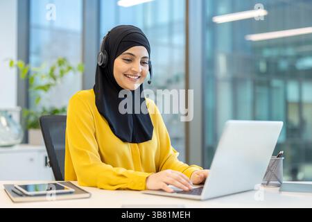 A smiling woman wearing a hijab and headset works at a desk, using a laptop in an office setting. The background features a window and office decor. Stock Photo