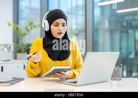 A woman wearing a hijab participates in a video conference, engaged in a discussion while seated at her desk. Stock Photo