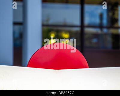A close-up view of a red circular object partially obscured by a white structure, set against a blurred background of windows and greenery. Stock Photo