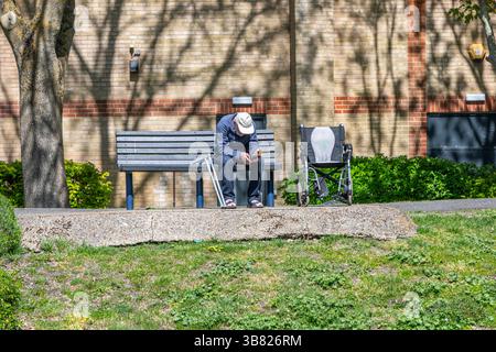 A solitary old man with a wheelchair and crutches sitting on a bench in the sunshine. Stock Photo