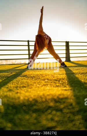 Curvy woman practicing yoga during golden hour sunset, stretching body in wide pose on green fitness platform near the sea, promoting wellness and bal Stock Photo
