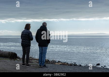 Rear view of two senior people enjoying the sea view in overcast cold day. Standing on the beach looking at the horizon over water. Unrecognizable act Stock Photo
