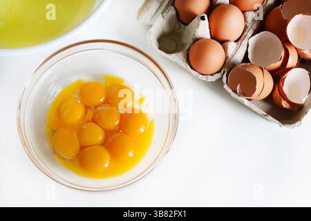 Separated egg yolks and whites in glass bowls and in the background broken eggshells and whole eggs. Directly above. Stock Photo