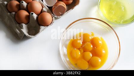 Separated egg yolks and whites in glass bowls and in the background broken eggshells and whole eggs. Directly above. Stock Photo