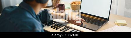 Man using MIDI keyboard and laptop for music production with coffee mug and cereals bowl on desk. Pressing keys and adjusting settings while enjoying Stock Photo