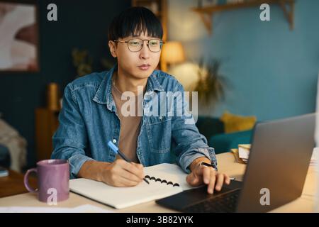 Portrait of young man with glasses sitting at desk, using laptop and writing in notebook showing concentration. Likely working or studying in cozy roo Stock Photo