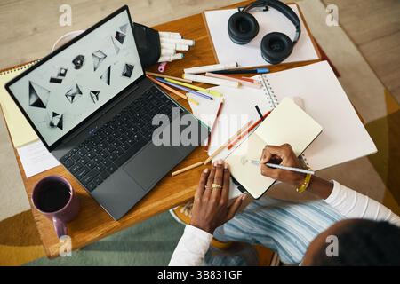 Woman working on creative project with laptop, notebook, and various stationery supplies on a wooden desk, including headphones, pens, and markers sur Stock Photo