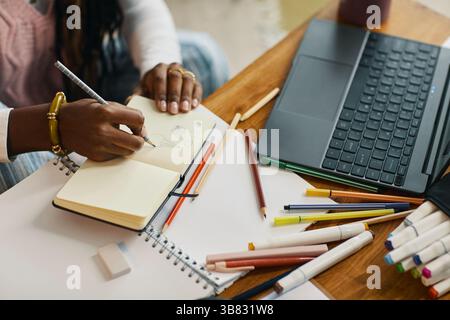 Close-up of woman writing notes in notepad while sitting at work desk with open laptop, pens, and pencils creating an organized workspace Stock Photo