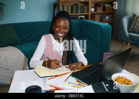 Smiling woman taking notes at desk filled with stationery items and laptop, looking comfortable and focused in cozy room, with bookshelves and seating Stock Photo