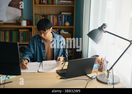 Man sitting at desk with multiple notebooks and a laptop, deeply focused on studying in home office filled with books and a lamp brightening workspace Stock Photo