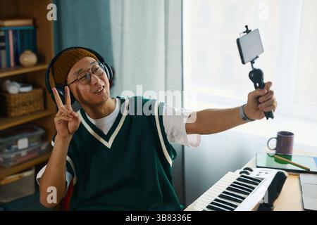 Man holding smartphone on extendable stick while gesturing peace sign and smiling. Wearing casual outfit and headset, sitting at desk in cozy room, sh Stock Photo