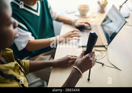 Multiracial team collaborating on project, using technology in office. Detail of coworker holding device while another uses laptop in background Stock Photo