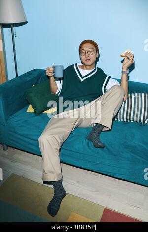 Young Asian man sitting comfortably on sofa, enjoying cup of tea and reading book. Room has modern furnishings including pillows and lamp in backgroun Stock Photo