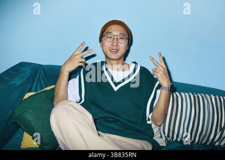 Portrait of young Asian man sitting on green couch wearing glasses and making peace signs with both hands wearing casual clothes and looking at camera Stock Photo