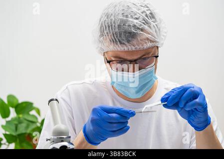 Scientific researcher wearing protective gear - blue gloves, surgical mask, hairnet, glasses - carefully handling laboratory sample near microscope using precision tweezers Stock Photo