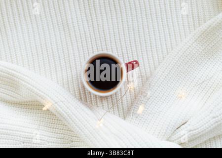 Warm coffee in a ceramic mug on a white knitted blanket with fairy lights. Cozy winter atmosphere, minimalistic holiday, Christmas new year scene. Stock Photo