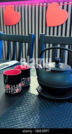 Elegant black teapot and artistic cups on a dark table setting in a romantic cafe with red heart decorations. Stock Photo