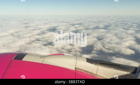 Vibrant pink airplane wing over a vast sea of soft white clouds under a clear blue sky. Captures the serenity and awe of in-flight travel. Stock Photo