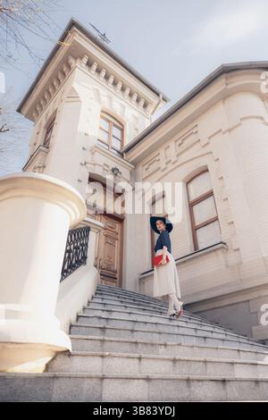 Elegant lady in hat walks up wide stone stairs with red book near white building in morning sun. Stock Photo