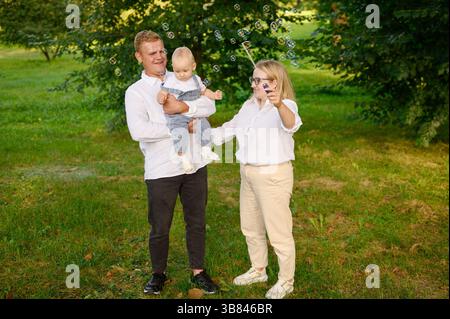 Family relaxing in park on weekend. Mom blowing soap bubbles, dad holding baby in arms, having fun, laughing. Family catching bubbles, playing togethe Stock Photo