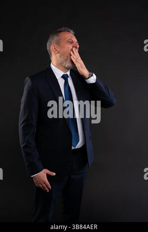 Full body portrait of a tired businessman in suit and tie, yawning and covering his mouth with hand. Studio shot on black background Stock Photo