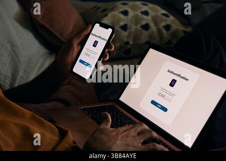 Midsection of man typing password on laptop for authentication through smart phone at home Stock Photo