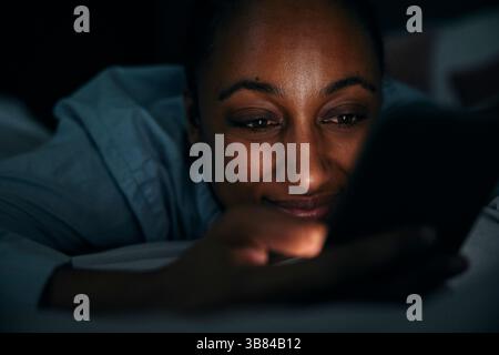 Smiling woman using smart phone while lying on bed in bedroom at night Stock Photo