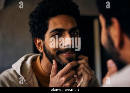 Smiling man with hand on beard looking at mirror in bathroom Stock Photo