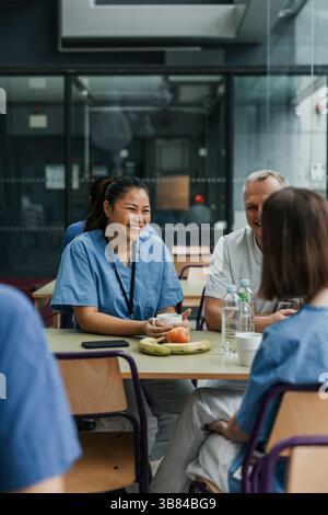 Happy young female medical professional having coffee while talking and sitting with colleagues at table in hospital caf Stock Photo