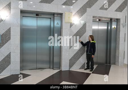 11 year old boy pressing an elevator button in a modern building lobby with stainless steel doors and geometric tile walls Stock Photo