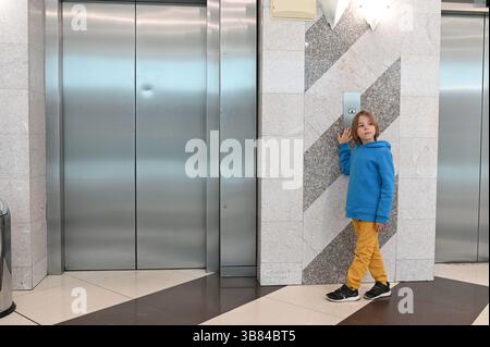 7 year-old boy in a blue hoodie pressing the elevator button in a modern building lobby with stainless steel doors and patterned walls Stock Photo