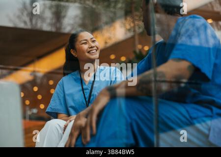 Happy female healthcare professional talking with male coworker while sitting in hospital building seen through glass Stock Photo