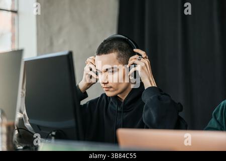 Focused non-binary entrepreneur wearing wireless headphones while working on desktop PC in office Stock Photo