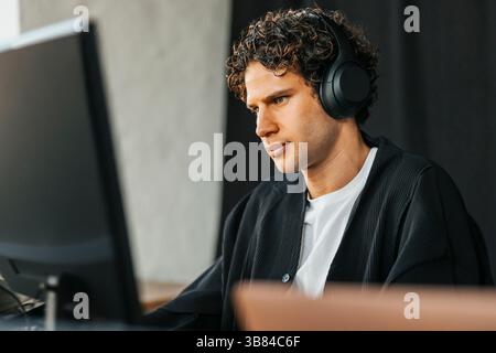Curly haired young male computer programmer wearing wireless headphones using desktop PC in startup office Stock Photo