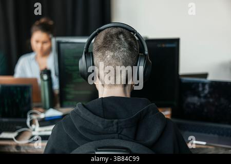 Rear view of non-binary IT professional wearing wireless headphones coding on desktop PC while working in office Stock Photo