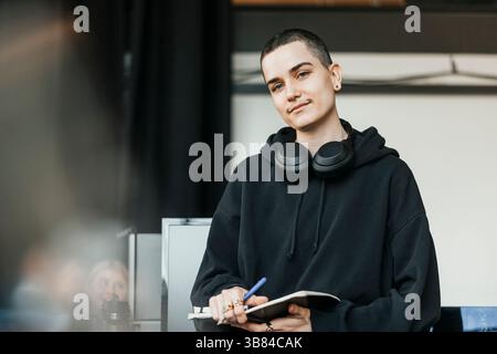 Portrait of confident non-binary computer programmer with wireless headphones around neck holding diary in hand Stock Photo