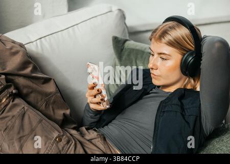High angle view of blond IT professional using smart phone while wearing wireless headphones lying on sofa in office Stock Photo