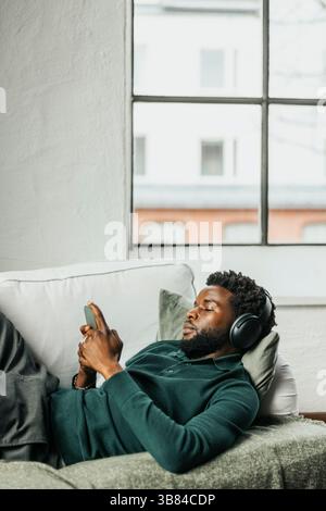 Relaxed businessman using smart phone while lying on sofa wearing wireless headphones in office Stock Photo
