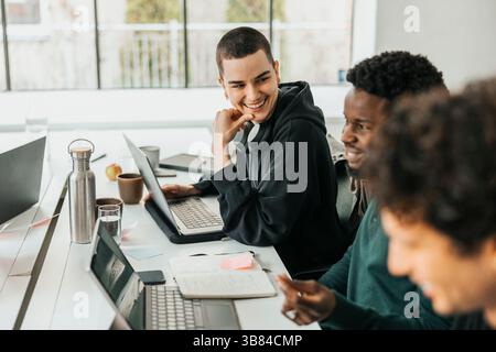 Happy non-binary software engineer sitting at desk while talking with colleague  in tech startup company Stock Photo