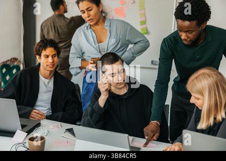 High angle view of young businessman sticking adhesive note on desk while planning strategies with colleagues in office Stock Photo