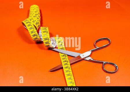 Top view of tailor dress measuring tape placed near sharp stainless steel scissors against orange background in light Stock Photo
