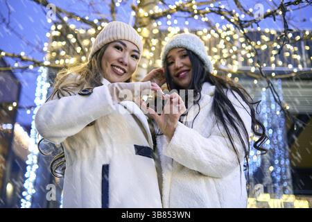 Two girlfriends asian girls in white coats are having fun, showing heart, hugging, new year, light bulbs Stock Photo