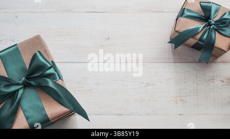 Two gift boxes in craft wrapping paper and green satin ribbon on white wooden table, copy space in center. Beautiful Christmas, New Year or Birthday p Stock Photo