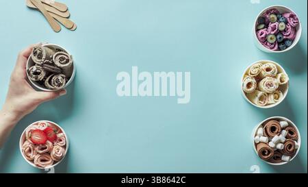 Hand with rolled ice cream in cone cup on blue background. Different iced rolls top view or flat lay. Thai style rolled ice cream with copy space in c Stock Photo