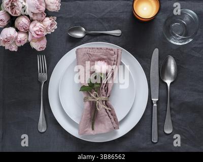 Beautiful table setting with candle on gray linen tablecloth. Festive table setting for wedding dinner with pink spray rose and pink napkin on plate. Stock Photo