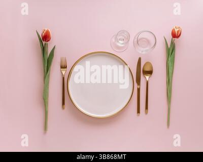 Festive pink table setting. Pink plate with gold cutlery and red tulip on pink background. Aesthetic stylish dinner table setting for Women's day, Mot Stock Photo