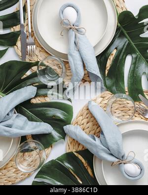 Tropical Easter Tablesetting. Top view of table setting for Christian Easter with bunny eggs decorated monstera leaves. Aesthetic Easter tablescapes. Stock Photo